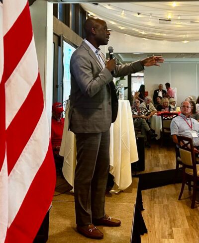 Berny Jacques addresses a room of activists. An American flag is in the foreground of the image.