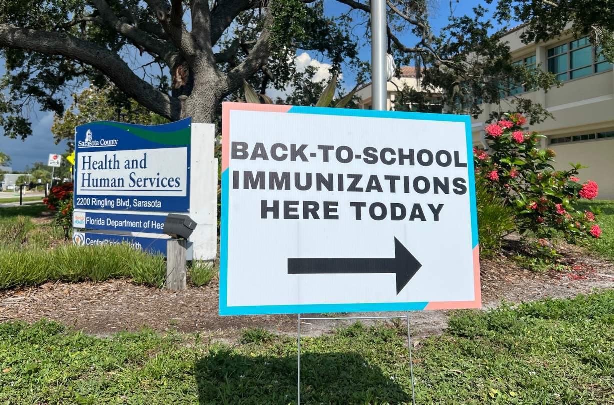 A yard sign in the foreground reads "BACK-TO-SCHOOL IMMUNIZATIONS HERE TODAY" with an arrow pointing toward a building. A sign in the background reads "Sarasota County Health and Human Services."