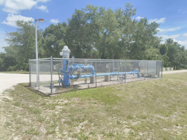 Blue pipes and machinery sit on grass, surrounded by a chain link fence.