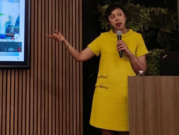 A woman speaks into a microphone while standing at a podium in front of a slideshow presentation