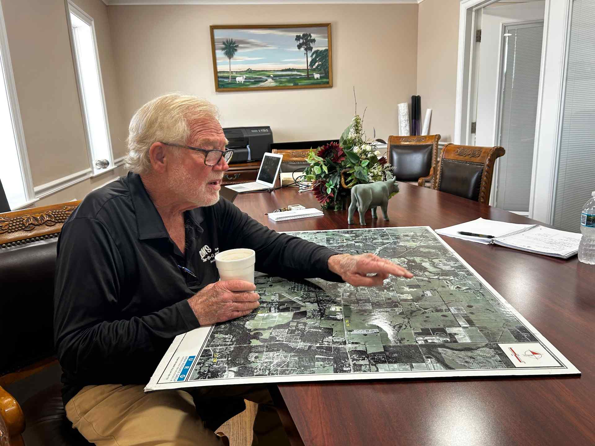 White haired man holding a coffee cup points at map spread across table in front of him