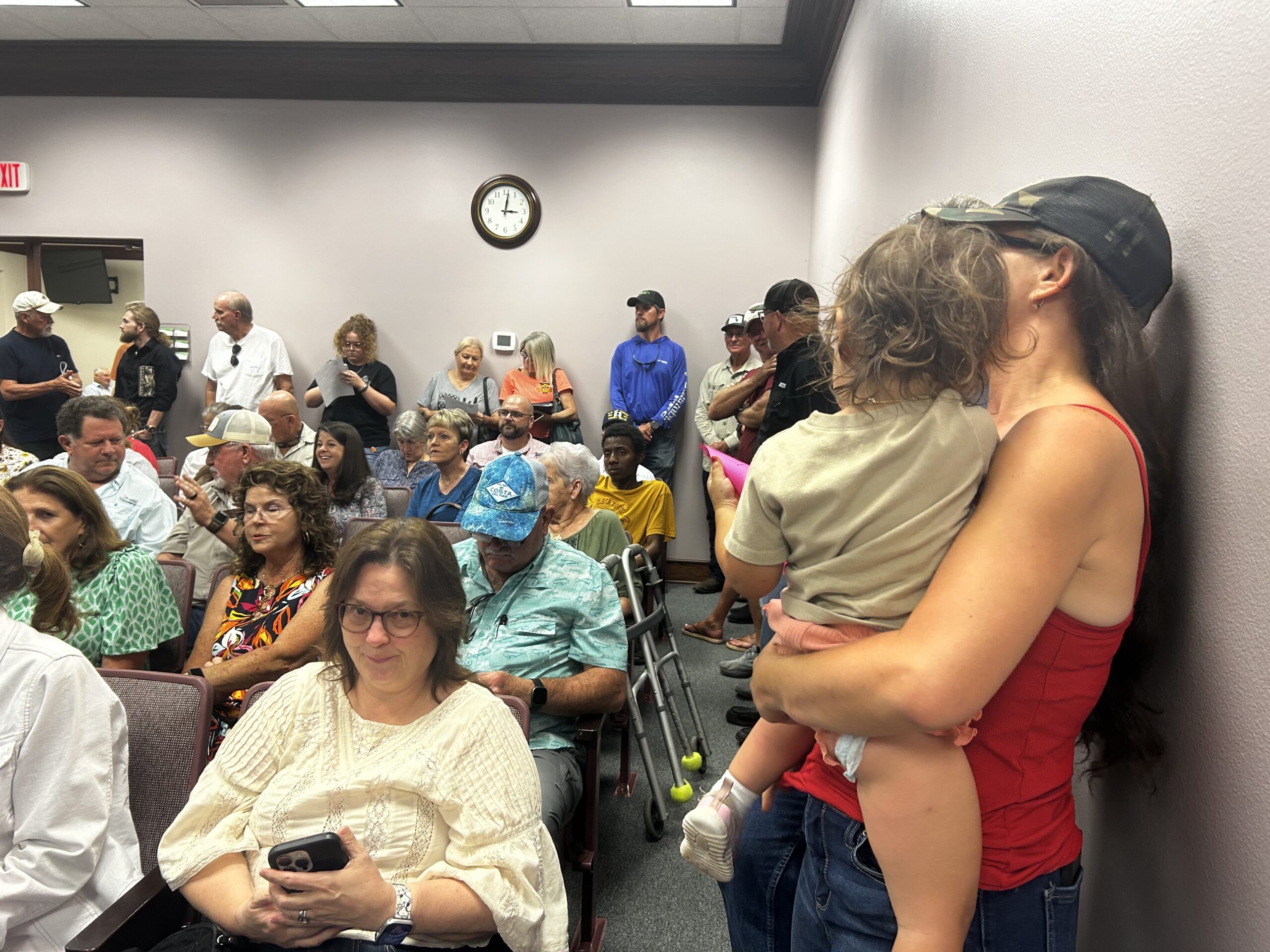 crowded room with people sitting in chairs and standing along the wall. mom and toddler are in foreground of photo.