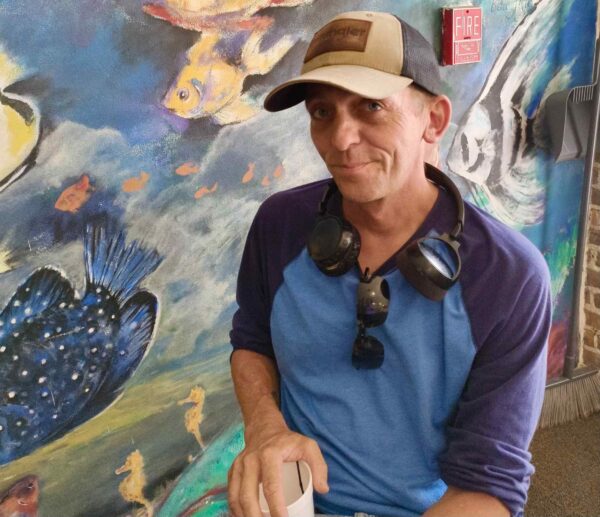 A man sits facing the camera in front of an ocean mural.