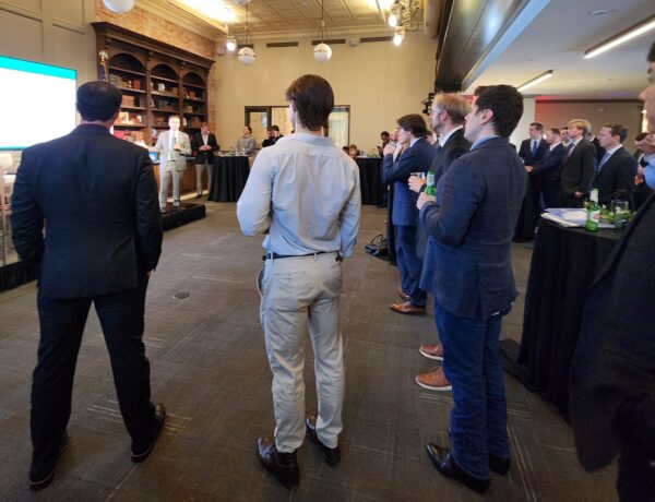 Men wearing suits stand facing away from the camera. They are watching a panel of speakers.