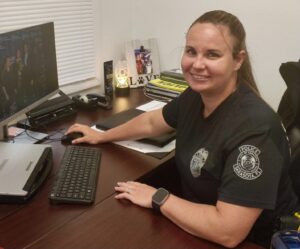 A woman in a police uniform smiles at the camera while sitting at a desk.
