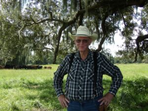 A man in a cowboy hat stands in a field with cows in the background.