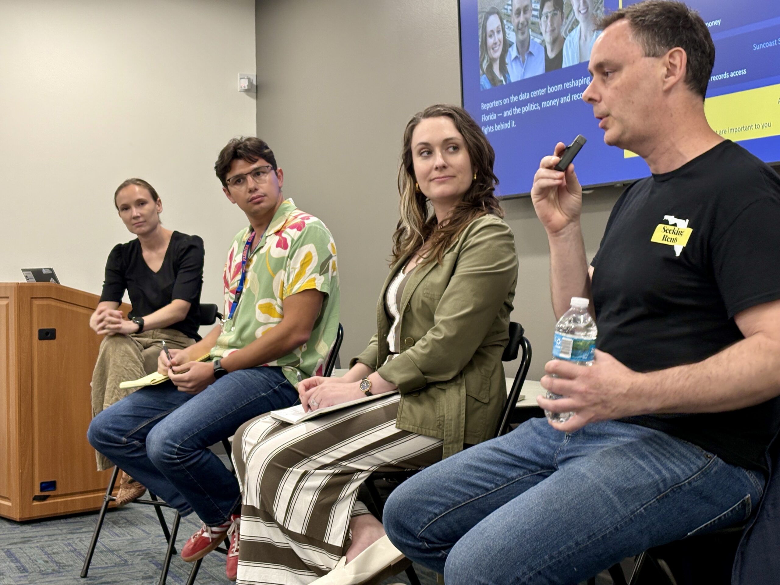 Panelists Four people sit in chairs at the front of a conference room.