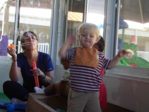 An adult sitting on the ground blows bubbles while toddlers flail their arms.