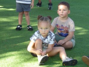 Two children sit on the ground. One is looking at the camera while tying her shoes, the other looks into the distance