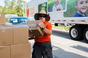 A man holds a box while standing next to a pile of other boxes. A semi-truck in the background says 