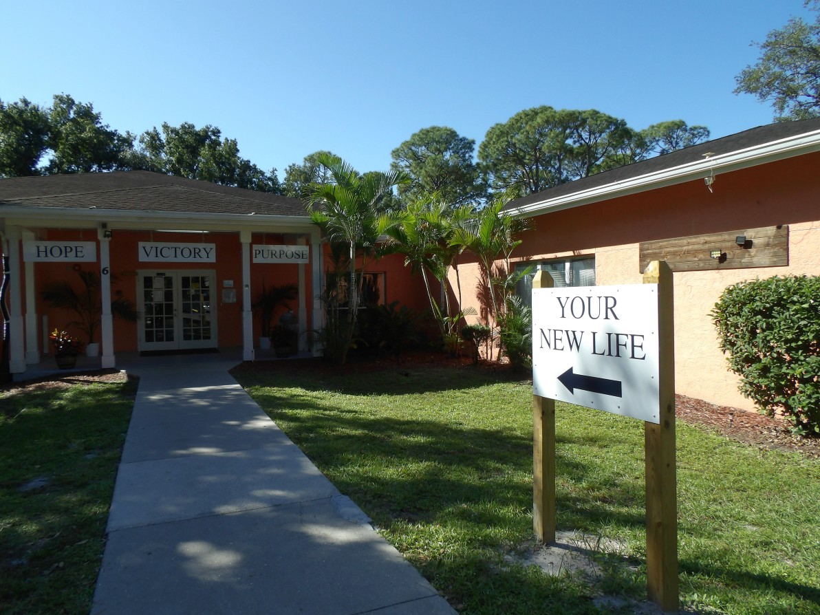 The Crossings entrance (1) A building entrance. Banners above the doors say "Hope", "Victory" and "Purpose." A sign on the path leading to the doors says "Your New Life" with an arrow pointing toward the building.